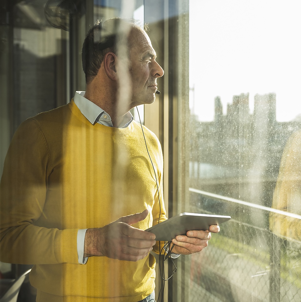 Contemplative businessman wearing headset standing with tablet PC seen through glass Unternehmer sieht nachdenklich aus dem Fenster